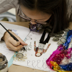 A student in an art class holds an ink pen while working on a drawing, which they lean over
