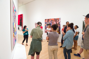 <p>A group of people, many with their arms crossed, stand in a white gallery with brightly colored works against the walls. A teaching artist in a teal shirt points towards the artwork behind her as she looks out to the group.</p>