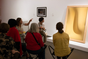 <p>Six people sit on stools in a white museum space. One person points toward a shadowy, yellow, abstract form in a wooden frame against the wall and the rest gaze towards it.</p>
