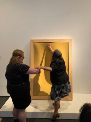 <p>A woman steps onto a white platform and reaches upward to touch a yellow foam compressed inside a wooden frame. Another woman stands on the ground, offering the assist of her elbow.</p>