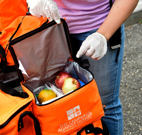 A person with gloved hands holds open an orange insulated bag containing fruit and other food items
