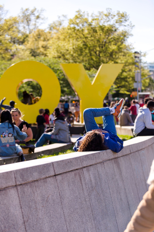 <p>A visitor lies on their back in the sun on the Museum plaza, with other people and the yellow Deborah Kass OY/YO sculpture in the background</p>