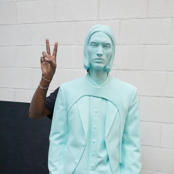 Virgil Abloh stands behind one of his works with only his arm and hand showing, making the peace/victory hand gesture