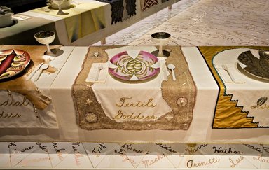 Judy Chicago (American, born 1939). <em>Fertile Goddess Place Setting</em>, 1974-1979. Runner:Cotton/linen base fabric, woven interface support material (horsehair, wool, and linen), cotton twill tape, silk, synthetic gold cord, flax warp, wool, hair weft, shells, bone needles, starfish, ceramic fetish figures, beads, coiled wool yarn medallions, wool cording, thread
Plate: Porcelain with overglaze enamel (China paint), rainbow luster, Runner:53 3/8 x 31 3/8 in. (135.6 x 79.7 cm). Brooklyn Museum, Gift of The Elizabeth A. Sackler Foundation, 2002.10-PS-2. © artist or artist's estate (Photo: , 2002.10-PS-02_Fertile_Goddess_Jook_Leung_photo_9286r2.jpg)